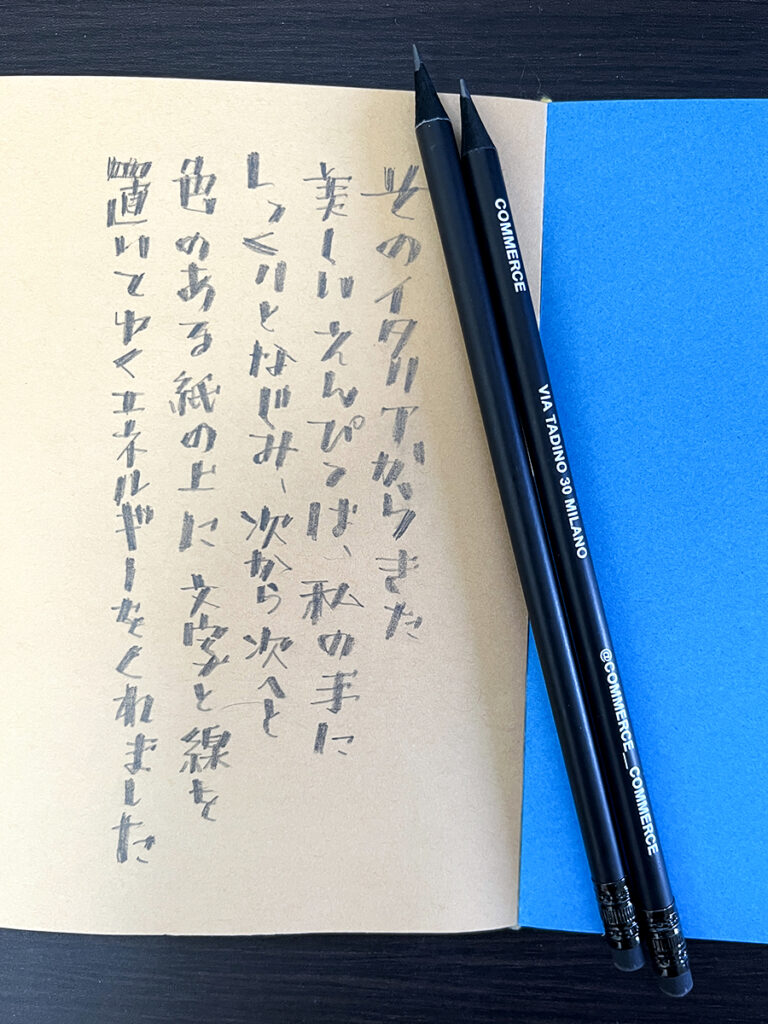 茶色い紙に鉛筆で書かれた日本語の縦書き文字と、2本の黒い鉛筆。イタリアの鉛筆で文字を描く喜びをテーマにしたカリグラフィー作品です。