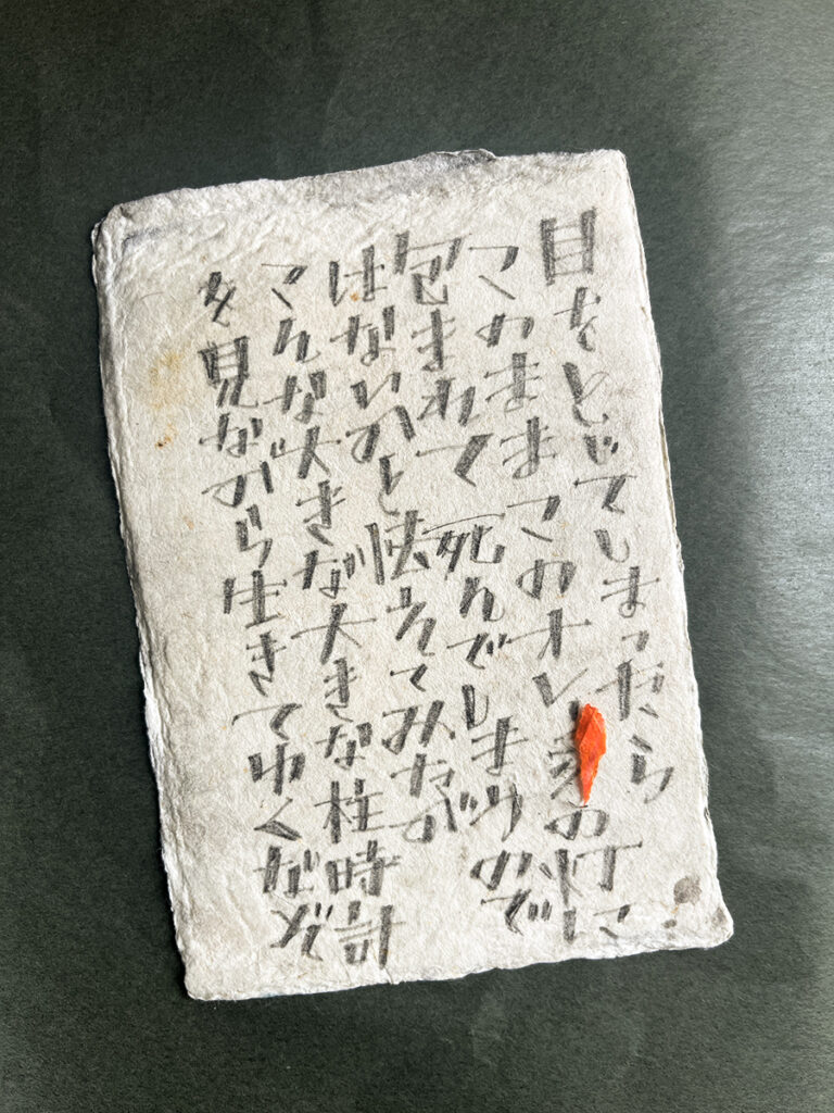 掠れた墨で縦書きされた文字と、右下に添えられたオレンジ色の紙片が印象的な、手漉き和紙のようなカリグラフィー作品。